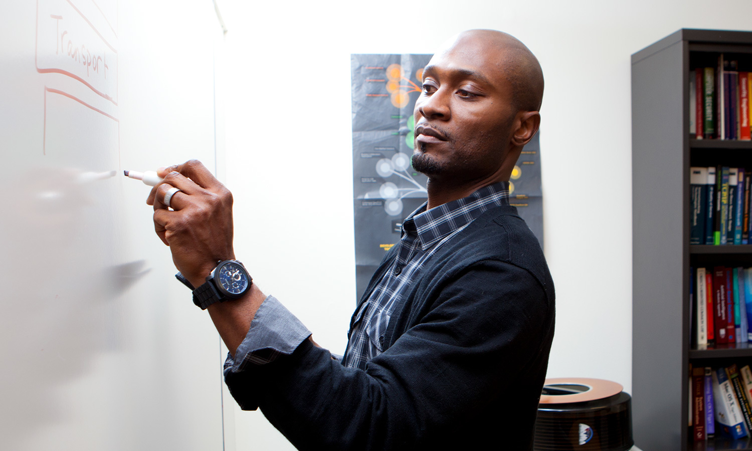 A man in writing on a whiteboard with focus with a bookshelf and scientific poster in the background.