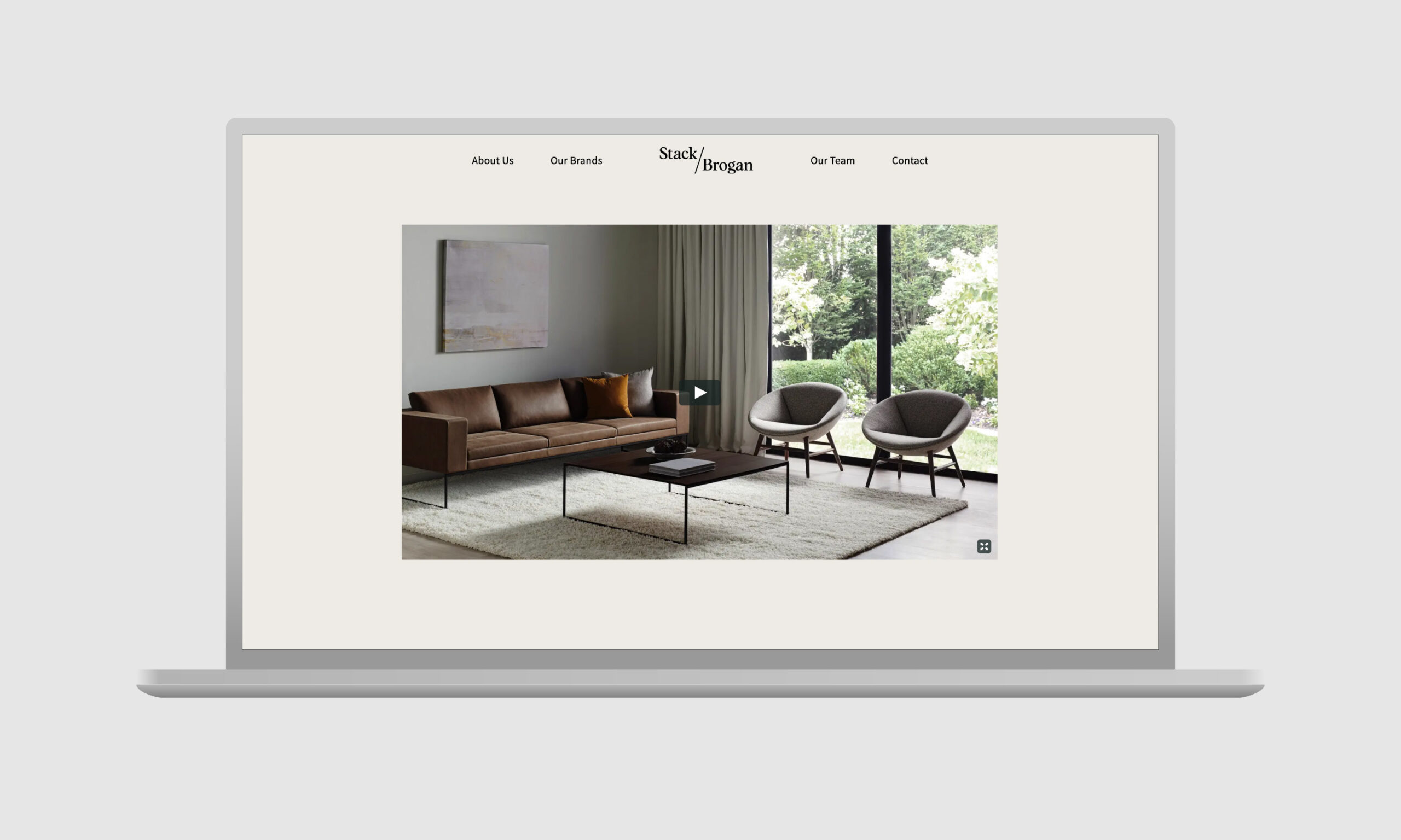 Laptop displaying the home page of the Stack Brogan website — a minimalist living room image with a brown sofa, two armchairs, and a coffee table on a rug. Large window shows greenery outside.