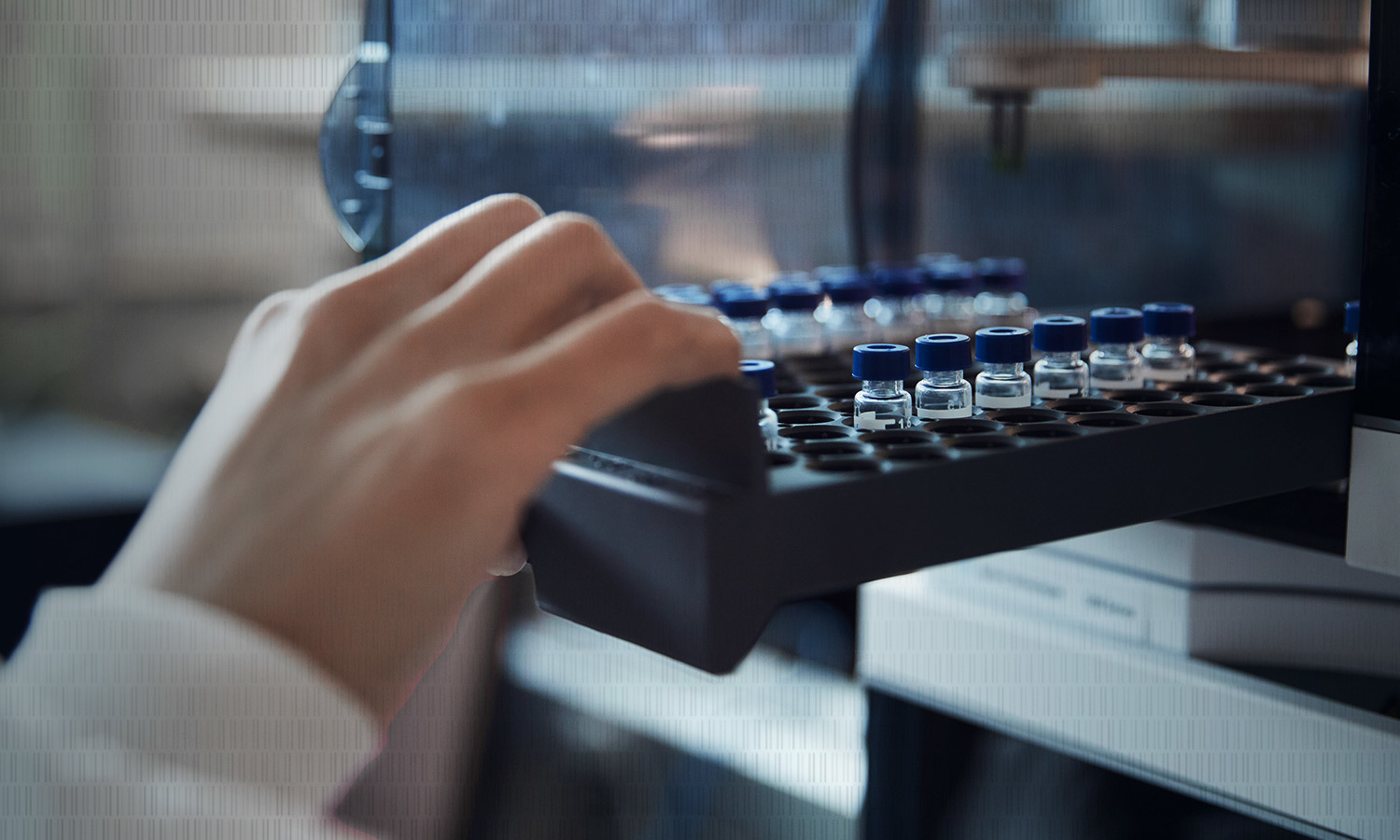 A person's hand places a tray filled with small, clear vials with blue lids into a laboratory machine.