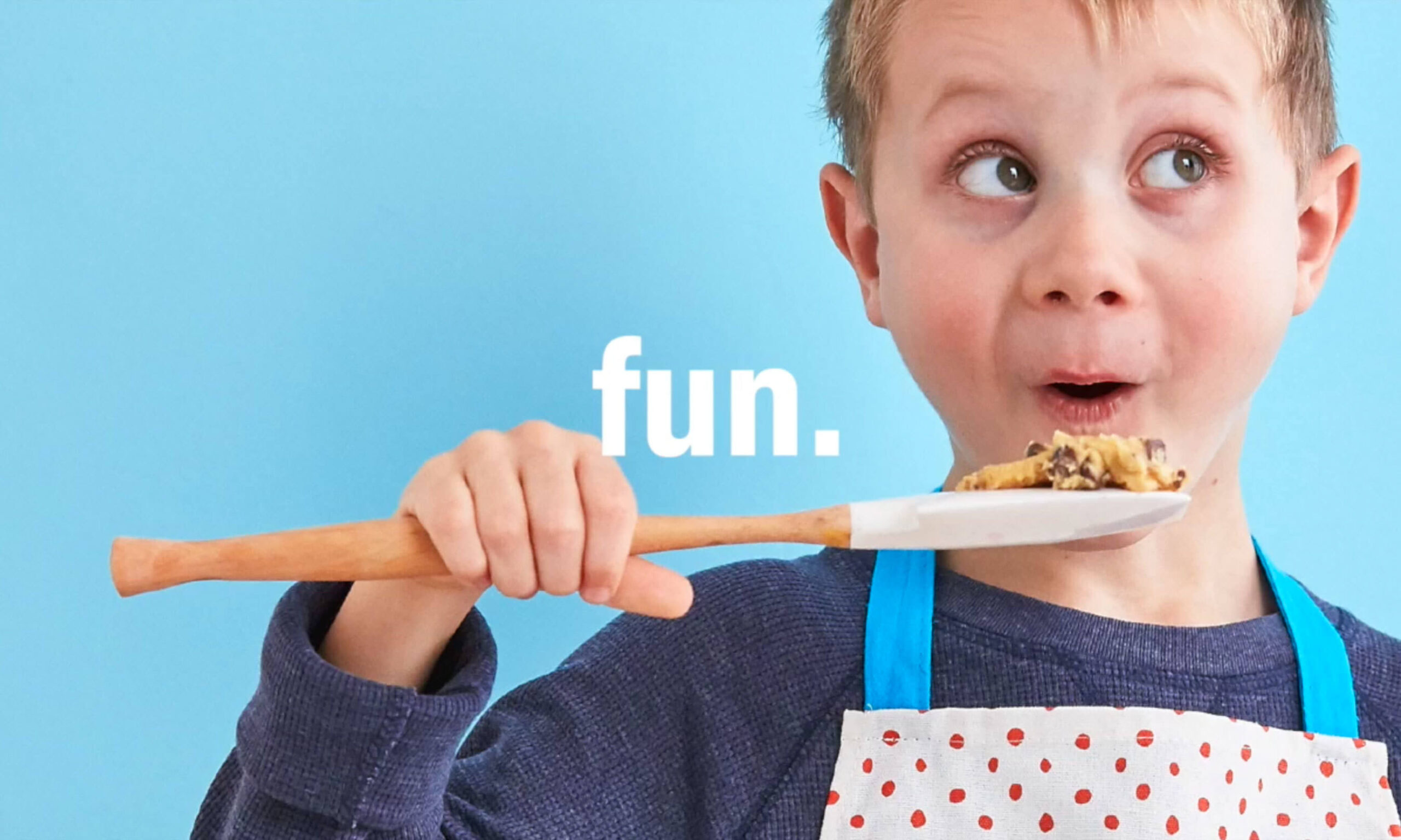 A young boy in a polka dot apron holds a spoon with cookie dough, looking surprised and playful. The word "fun" is centered against a blue background.