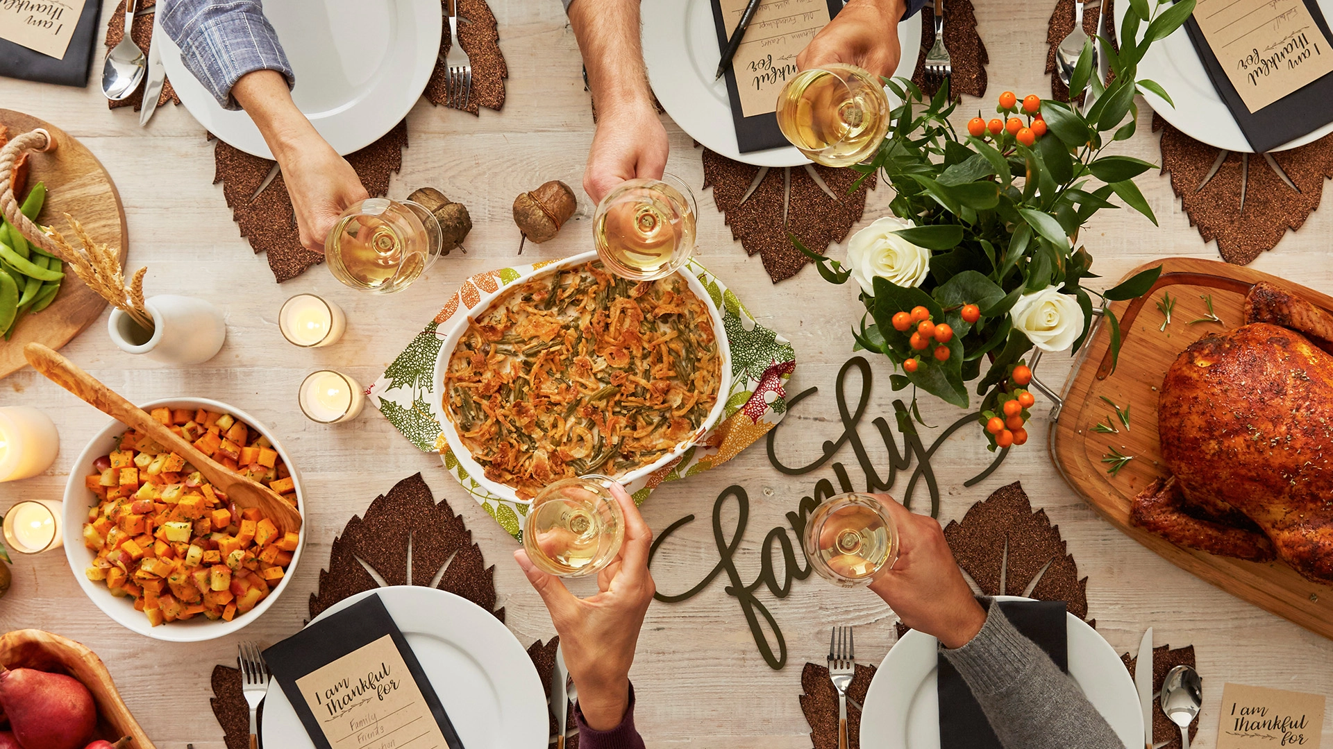 Festive table with roasted turkey, green bean casserole, and sweet potatoes. Six hands raise glasses, with flowers and a "family" script centerpiece. Warm, celebratory mood.