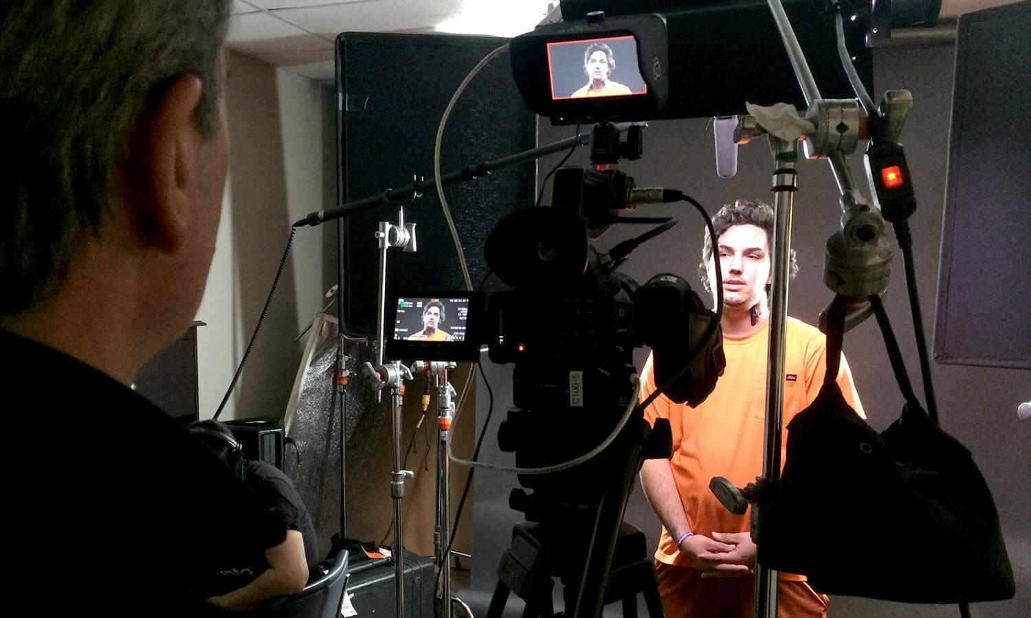 A person in an orange shirt is being filmed in a studio for the One20 video with professional camera equipment.